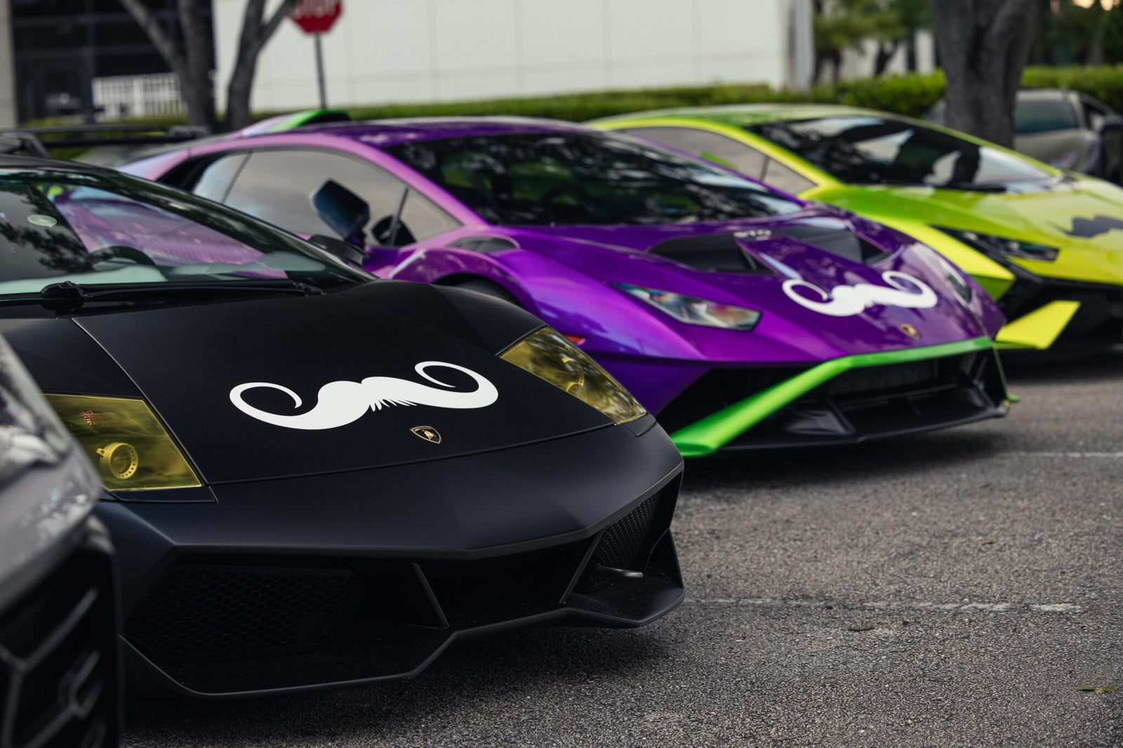 Three sleek sports cars, including a striking Lamborghini, are parked in a row, each proudly sporting a large mustache decal on the hood in support of Movember. The vibrant colors—black, purple, and green—make a bold statement for Men's Health awareness.