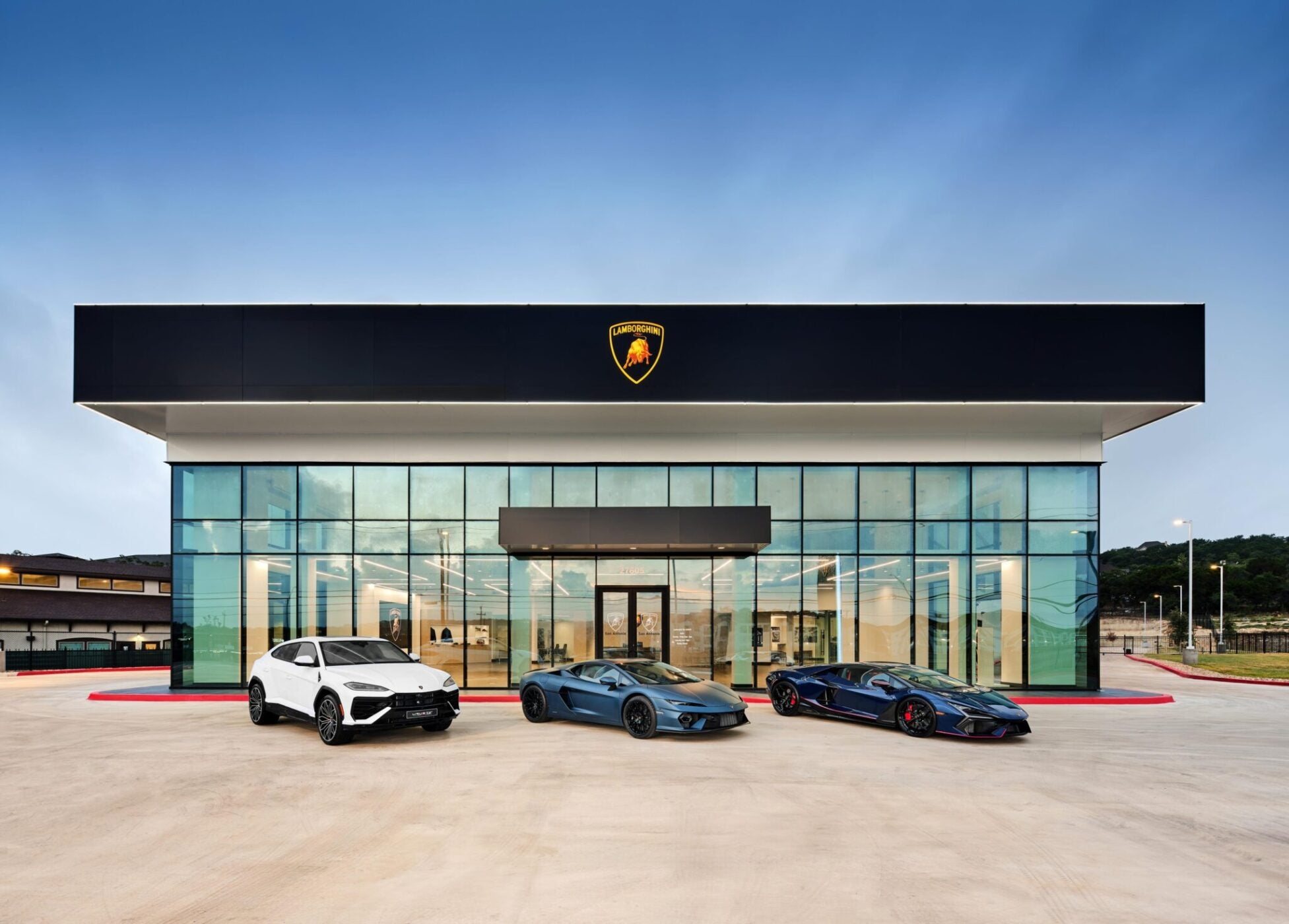 Three Lamborghini cars, part of the brand's record numbers, are parked in front of a modern dealership building with a glass facade under a clear blue sky.