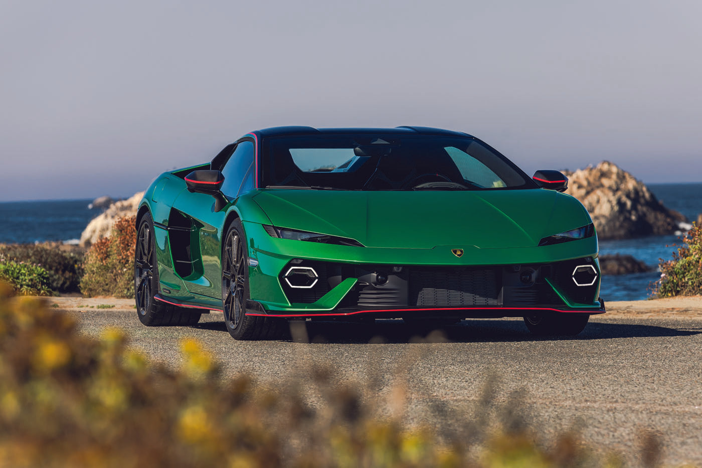 A sleek Lamborghini sports car, known as the Charging Bull, is parked on a coastal road with stunning ocean views and rugged rocks in the background.