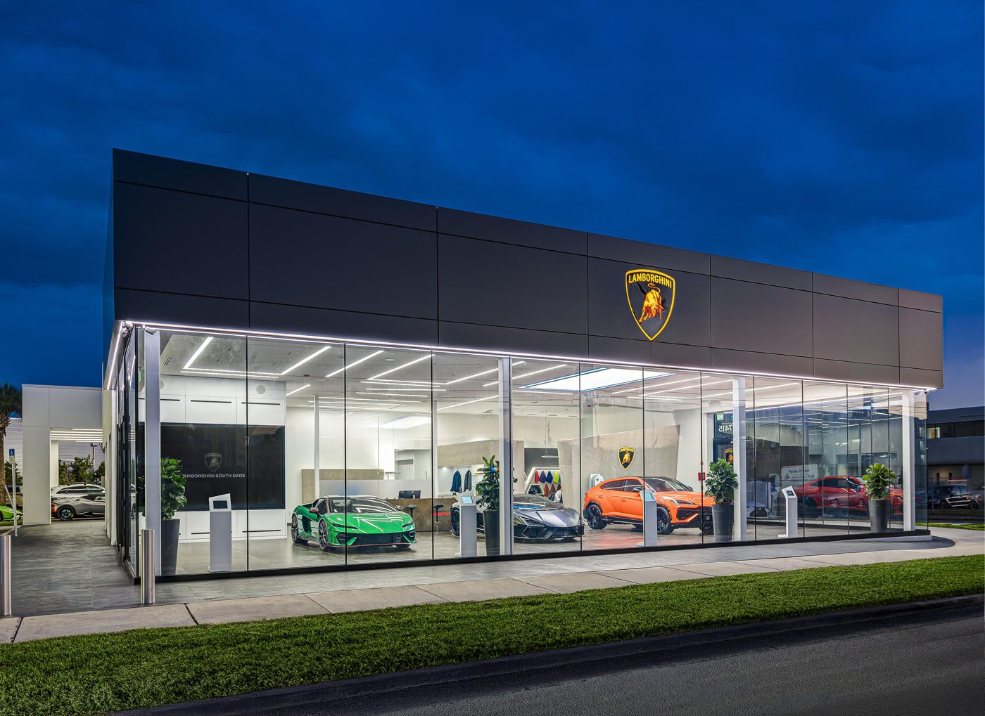 The Lamborghini showroom in South Dade glows at night, with an impressive lineup of cars displayed elegantly behind expansive glass windows.
