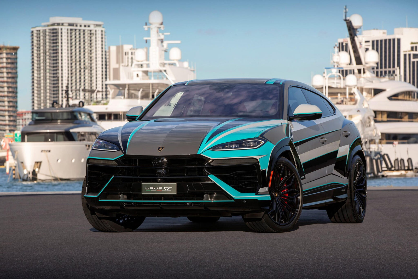 A black and turquoise Lamborghini Urus SE luxury SUV is parked near a marina, complemented by yachts and high-rise buildings in the background under a clear blue sky.