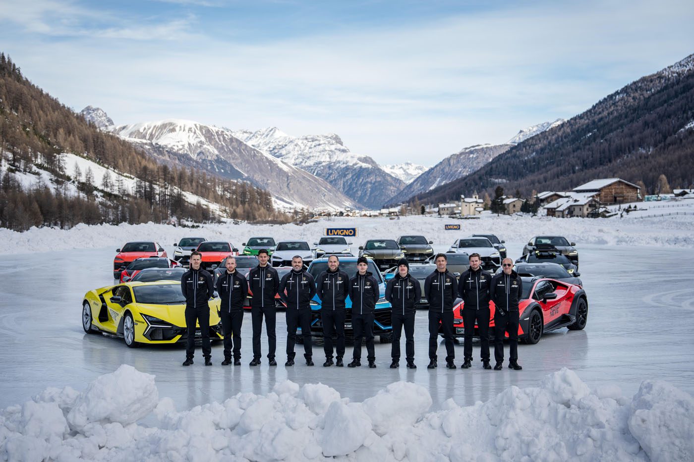 A group of twelve people wearing matching outfits stands on a snowy surface in Livigno, surrounded by sleek Lamborghinis against a stunning mountainous backdrop, setting the scene for an unforgettable snow driving experience.