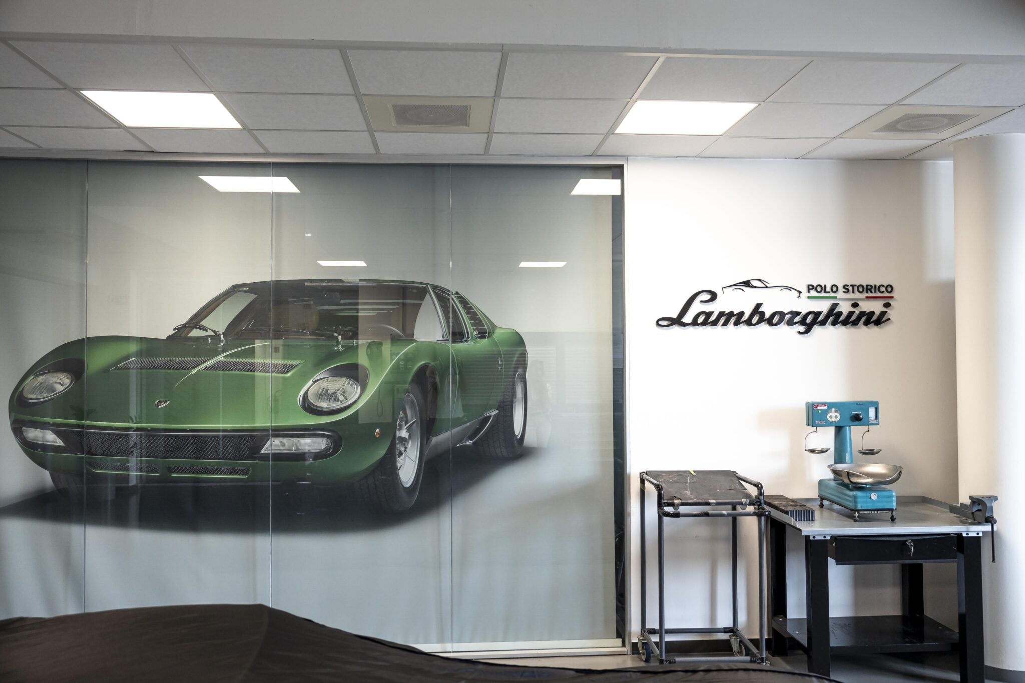 A green classic car is showcased behind glass, accompanied by "Polo Storico Lamborghini" signage, emblematic of a global celebration. To the right, a vintage machine rests on a small table.