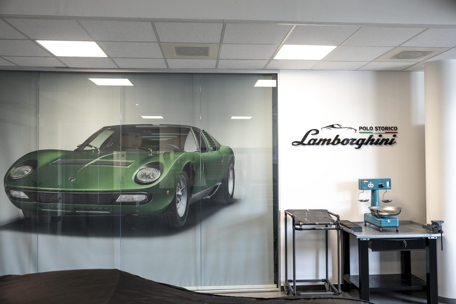 A green classic car is showcased behind glass, accompanied by "Polo Storico Lamborghini" signage, emblematic of a global celebration. To the right, a vintage machine rests on a small table.