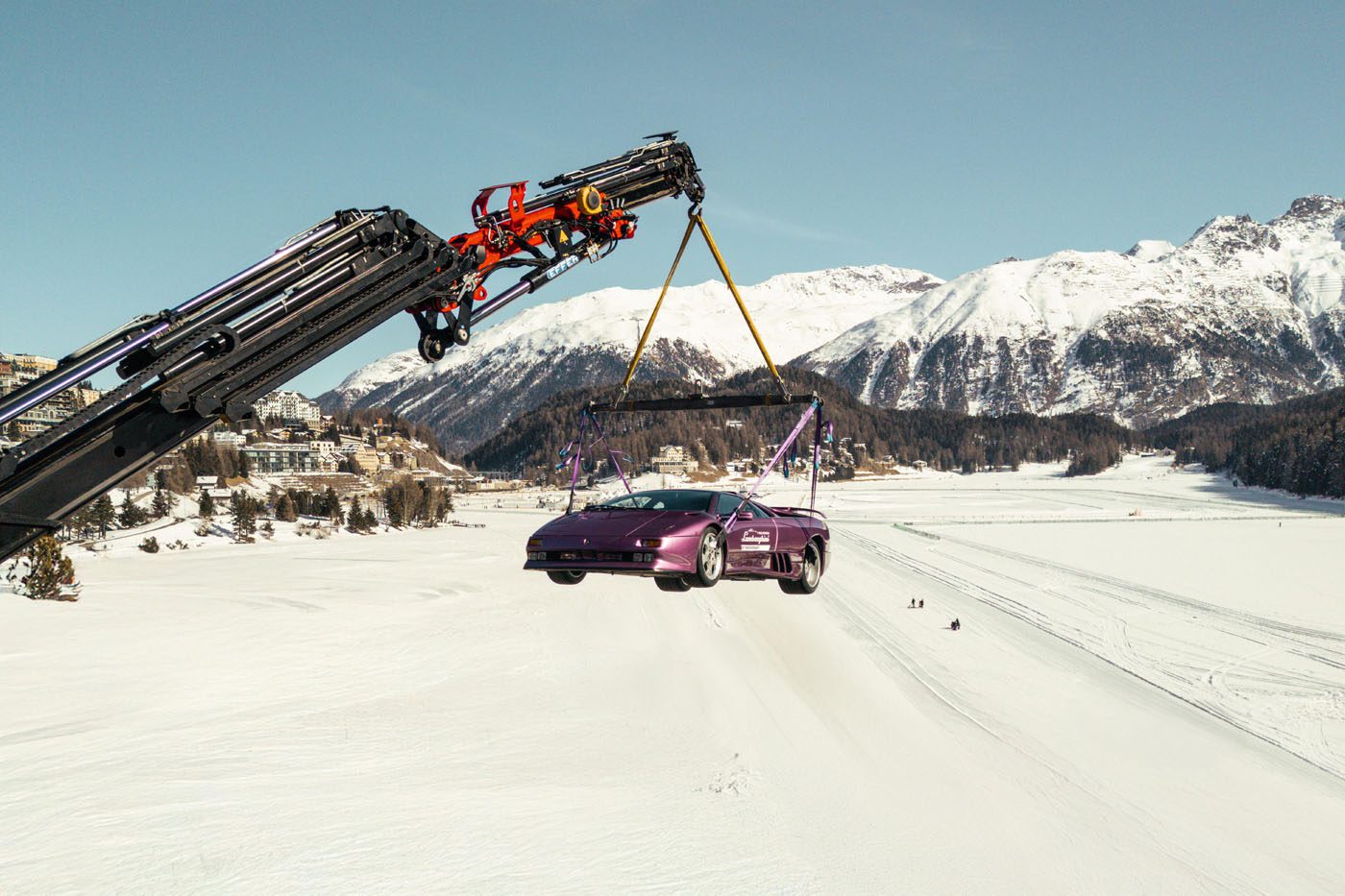 A Lamborghini is suspended by a crane over a snowy landscape, with mountains and buildings in the background, evoking the elegance of I.C.E. St. Moritz.