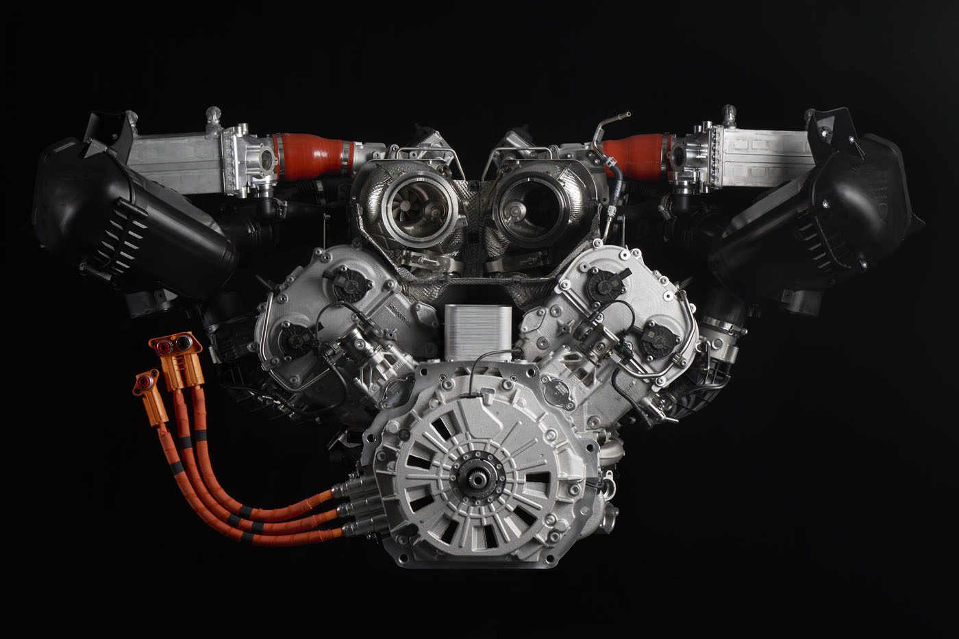 Close-up of a complex Lamborghini engine against a black background, featuring multiple components, orange cables, and metal parts symmetrically arranged within its 900HP powertrain.