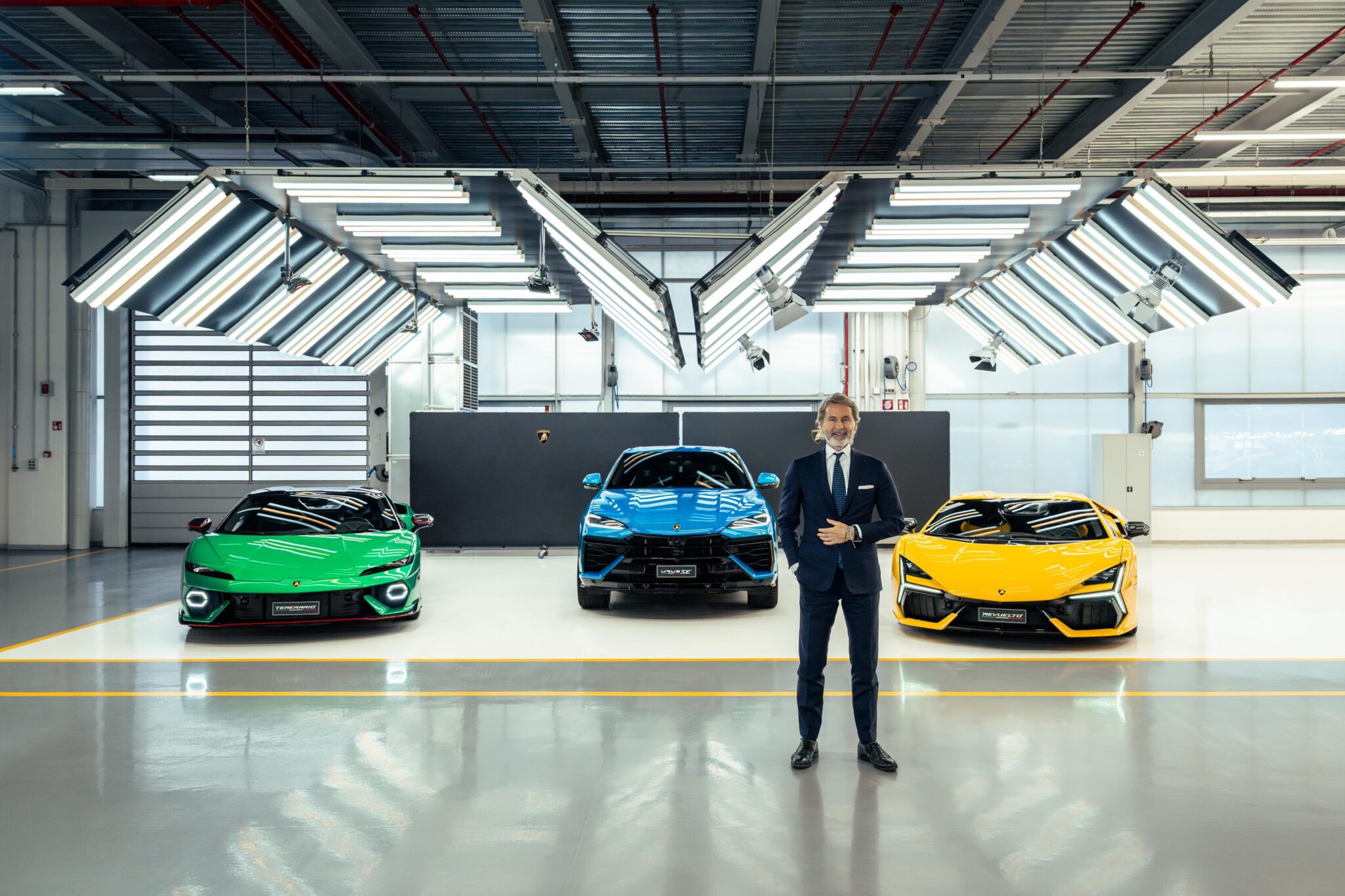 A man in a suit stands in front of three colorful sports cars—green, blue, and yellow Lamborghinis—inside a well-lit showroom with industrial ceiling lights, showcasing their anticipated 2024 sales.