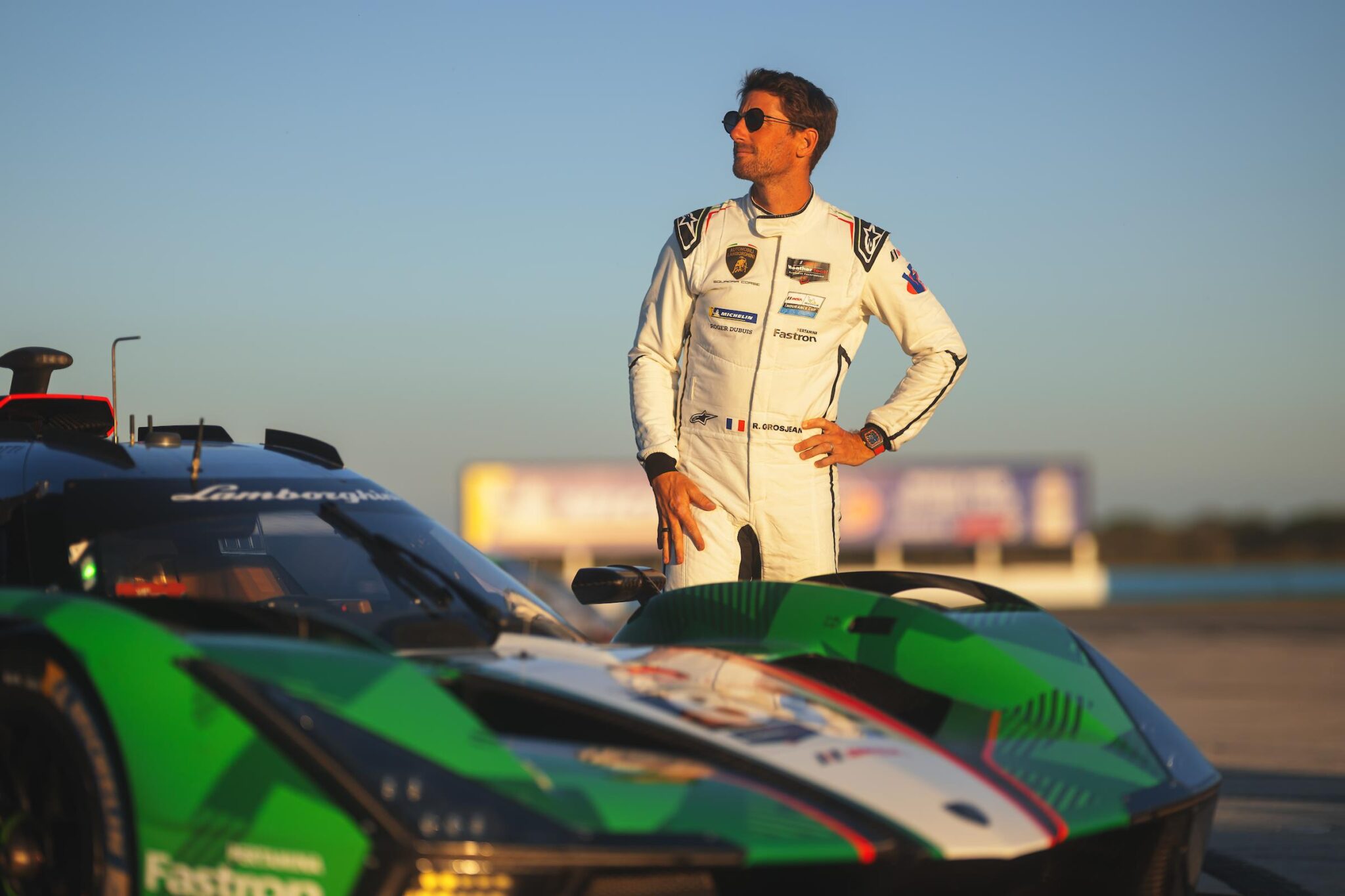Race car driver in sleek racewear by Alpinestars stands confidently beside a green and black Lamborghini Squadra Corse sports car on a racetrack under a clear sky.