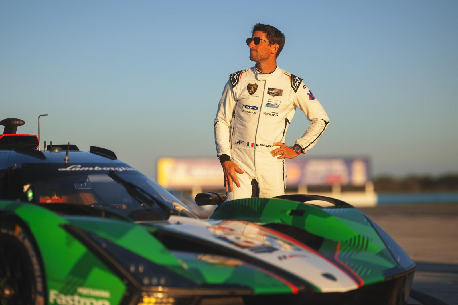 Race car driver in sleek racewear by Alpinestars stands confidently beside a green and black Lamborghini Squadra Corse sports car on a racetrack under a clear sky.
