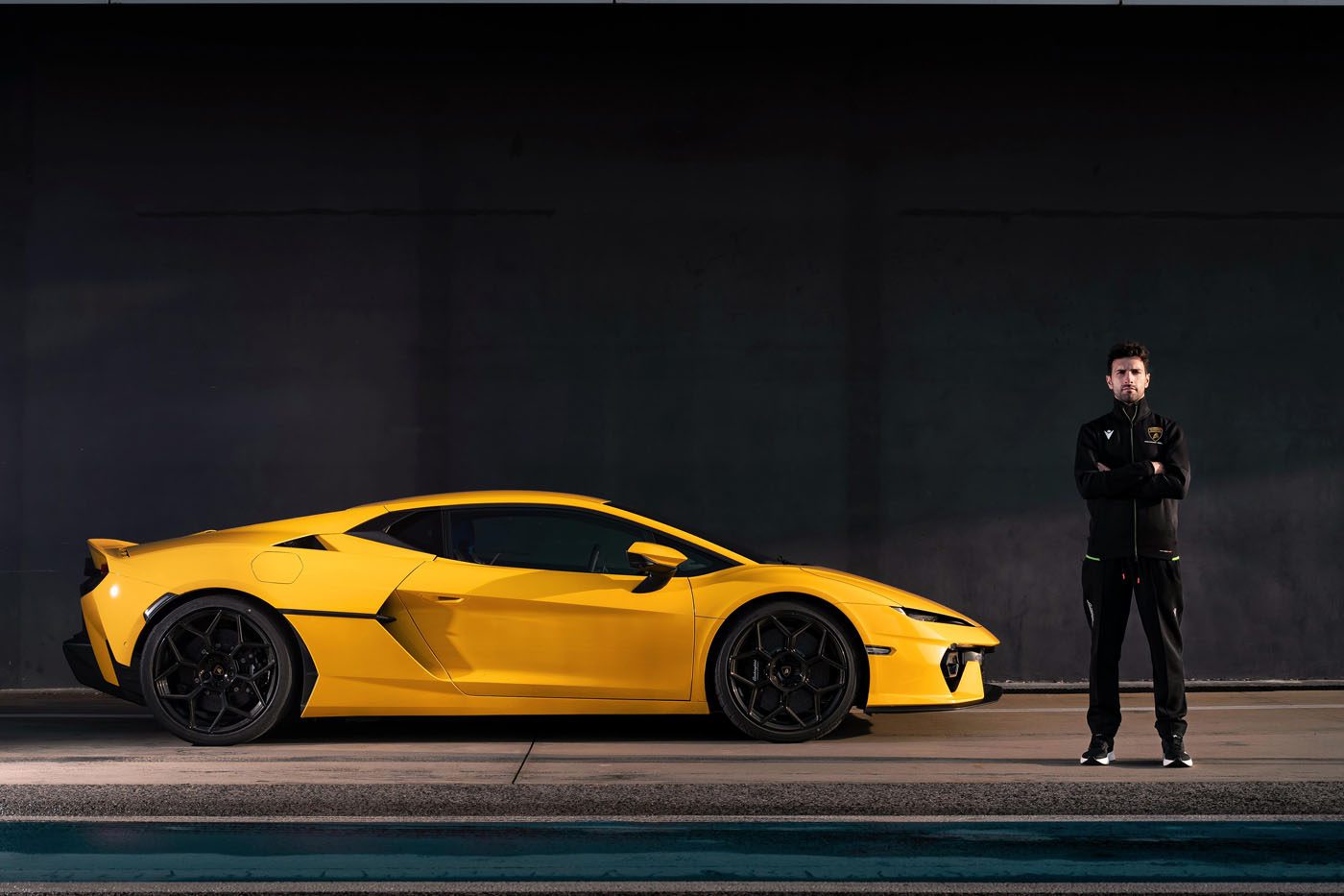 A person stands next to a sleek Lamborghini sports car in a dimly lit setting, exuding the aura of a factory driver ready to push limits.