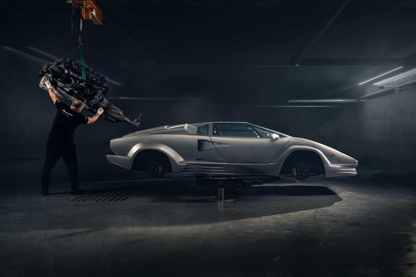A person uses a hoist to lift an engine near a wheel-less white Lamborghini Countach in an underground garage, possibly preparing for a Polo Storico restoration or display at an Anniversary Event.