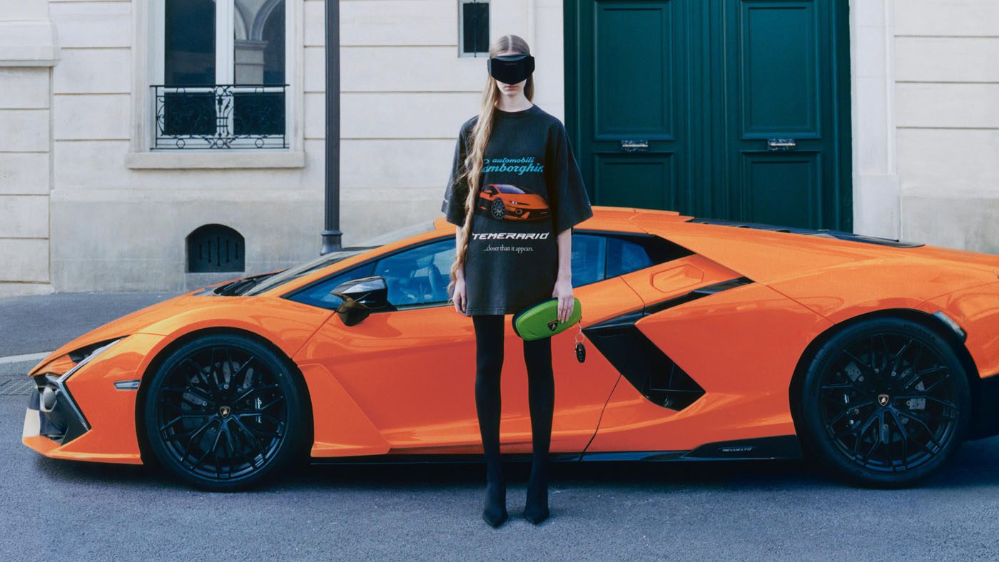 A person wearing black Balenciaga clothing, a visor, and holding a green clutch stands in front of an orange Lamborghini parked on a city street.