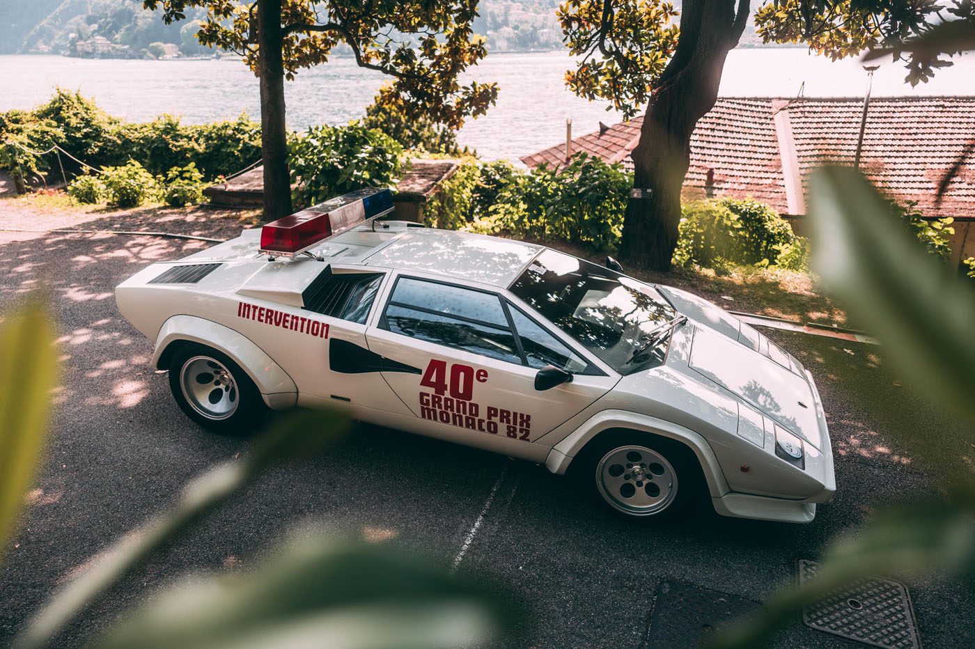 White Lamborghini Countach classic with "40e Grand Prix Monaco 82" and "Intervention" markings, parked outdoors near trees and a body of water, featuring a rooftop light bar—a true icon among race cars.