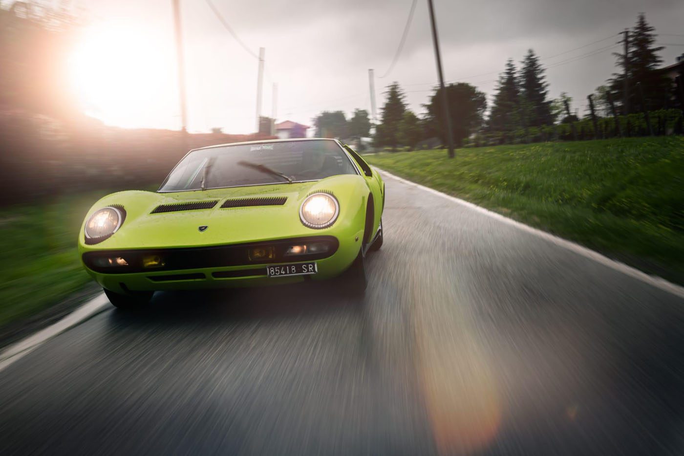 A green Lamborghini in stunning Verde Scandal cruises along a wet country road lined with trees and power lines, set against a cloudy sky—a story of speed and style.