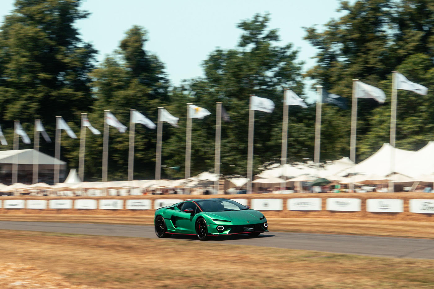 A green Lamborghini Temerario speeds along the track at Goodwood, with white flags and tents in the background, surrounded by trees.