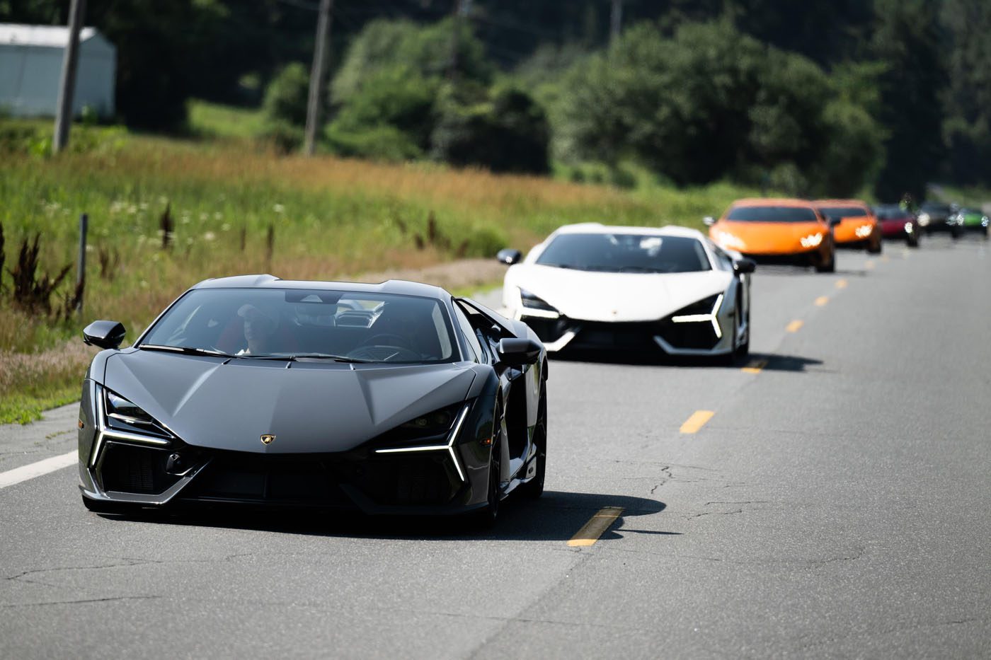 A line of luxury sports cars, led by a matte black Lamborghini, cruises down a two-lane road during the Esperienza Giro USA in New England, with trees and grass bathed in sunlight.