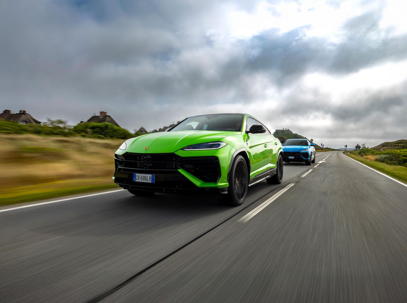 A bright green Lamborghini Urus SE leads a blue SUV on an empty, two-lane road near Sylt, with cloudy skies overhead and grassy shoulders—perfect for an exclusive event drive.
