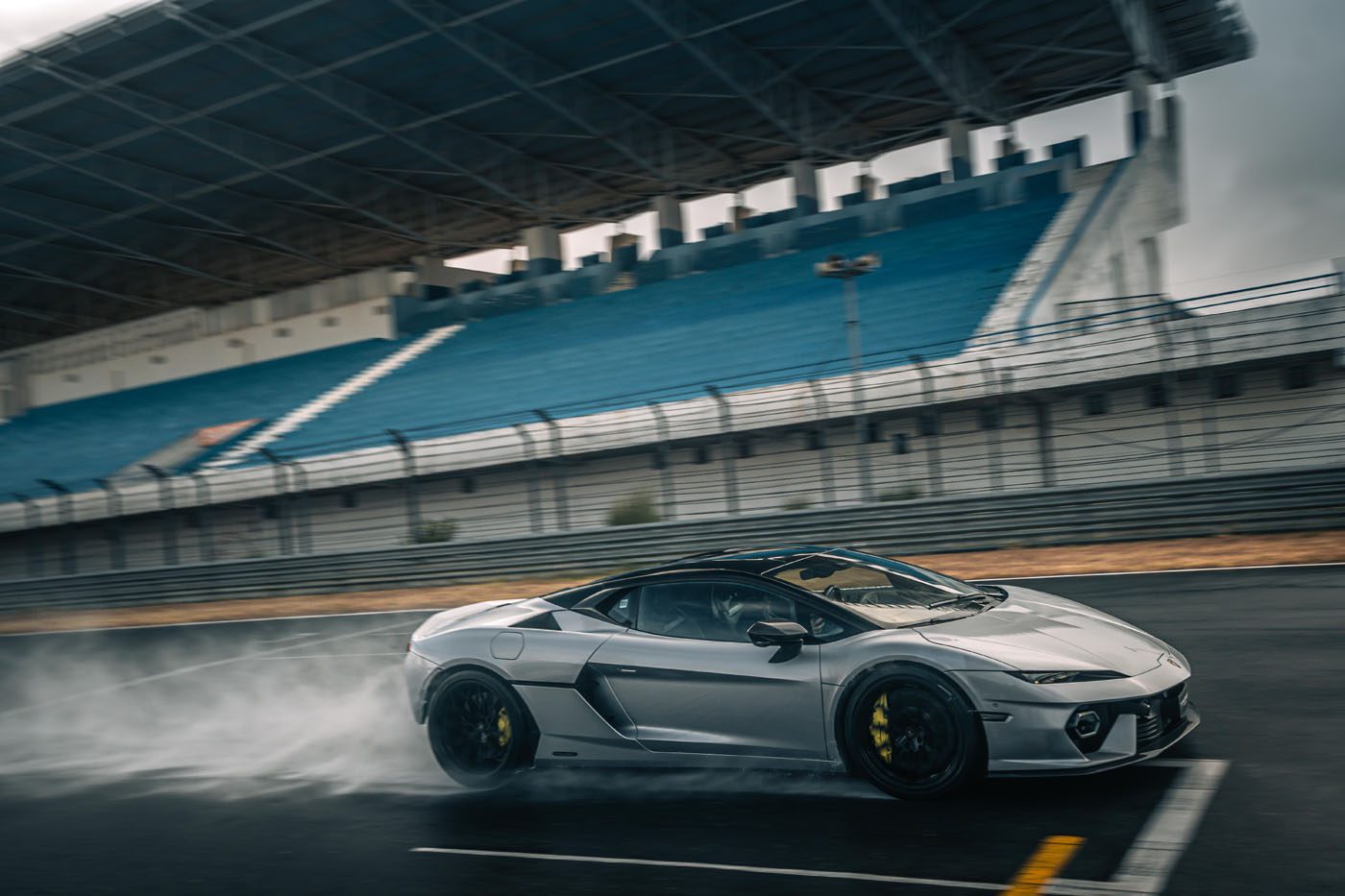 A silver Lamborghini Temerario makes a dynamic debut as it speeds on track, sending water spray flying near empty blue stadium seats under a large canopy.