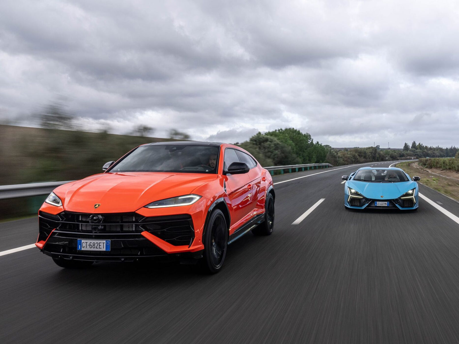 Two cars, an orange SUV and a blue Lamborghini sports car, drive side-by-side on a multi-lane highway under a cloudy sky.