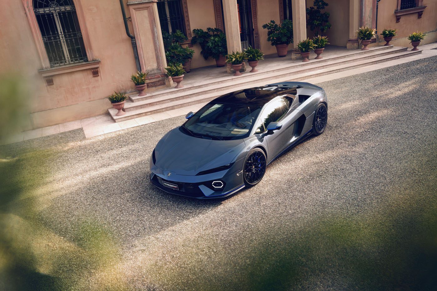 A grey Lamborghini Temerario is parked on a gravel driveway in Porto Cervo, in front of a building with columns, potted plants, and large windows.