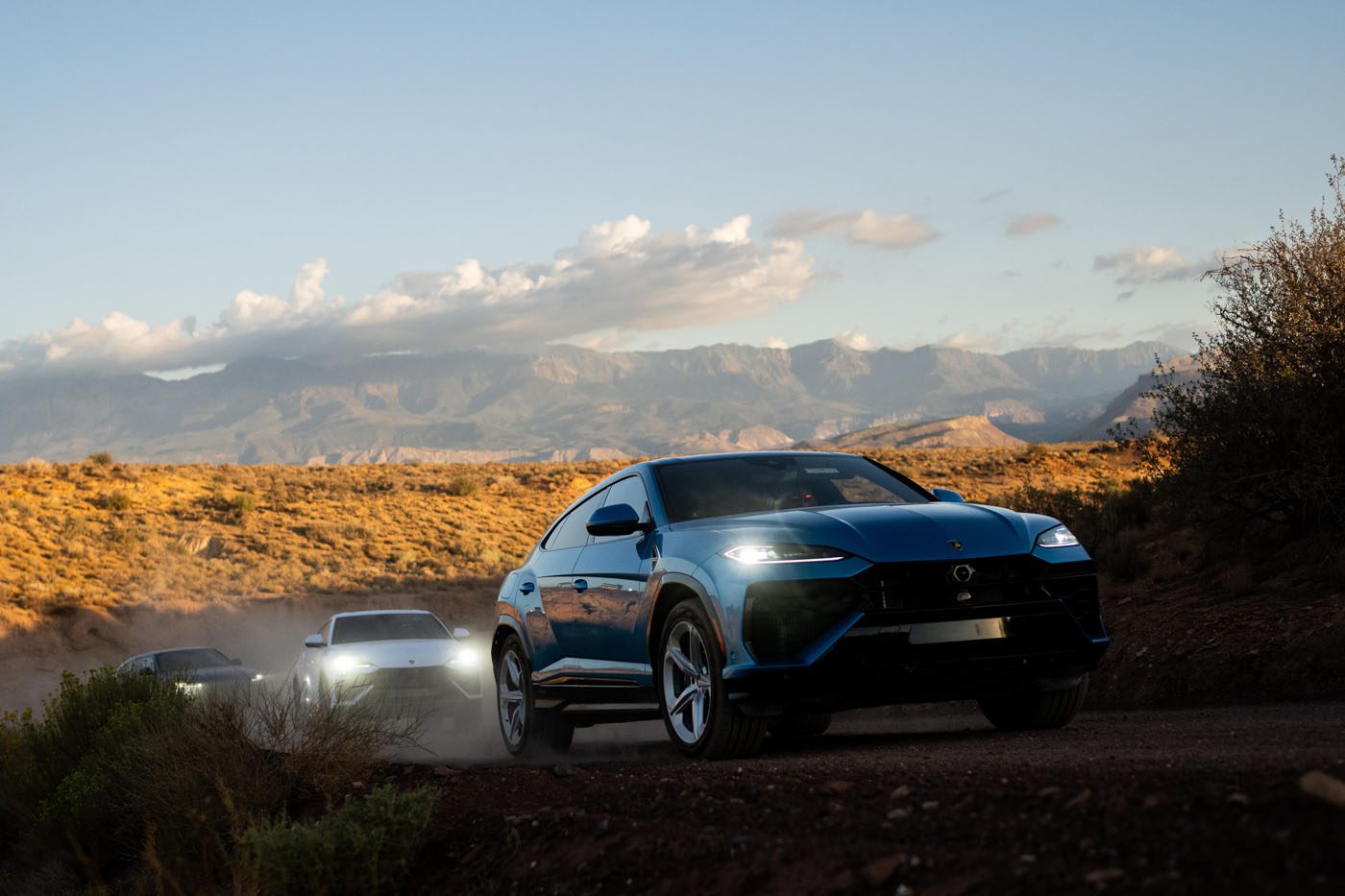 Three SUVs, including a Lamborghini Urus SE, drive on a dusty dirt road through a desert landscape near Zion National Park, with mountains and clouds in the background.
