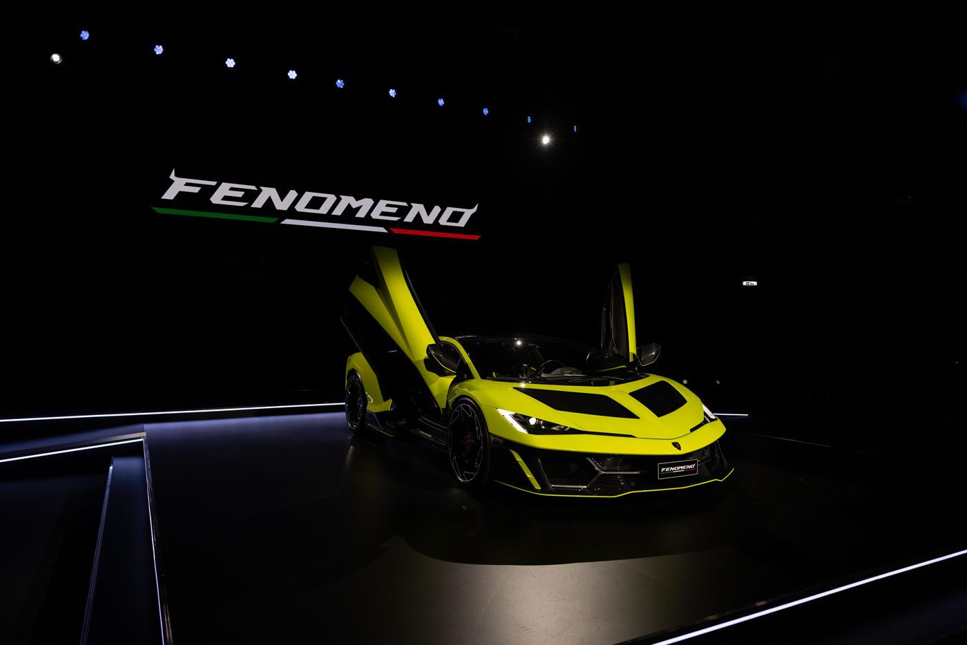 A bright yellow sports car with scissor doors open is displayed on a dark stage beneath a sign that reads "FENOMEN," headlining the Fenomeno Display at Lamborghini Day UK.
