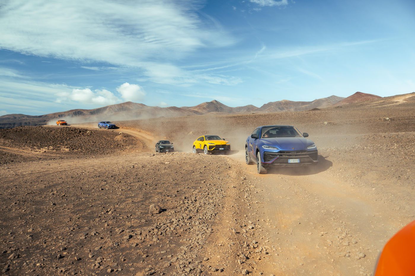 A convoy of colorful SUVs, including the Urus SE, drives on a dusty, rocky dirt road through an arid, mountainous Lanzarote landscape under a blue sky.