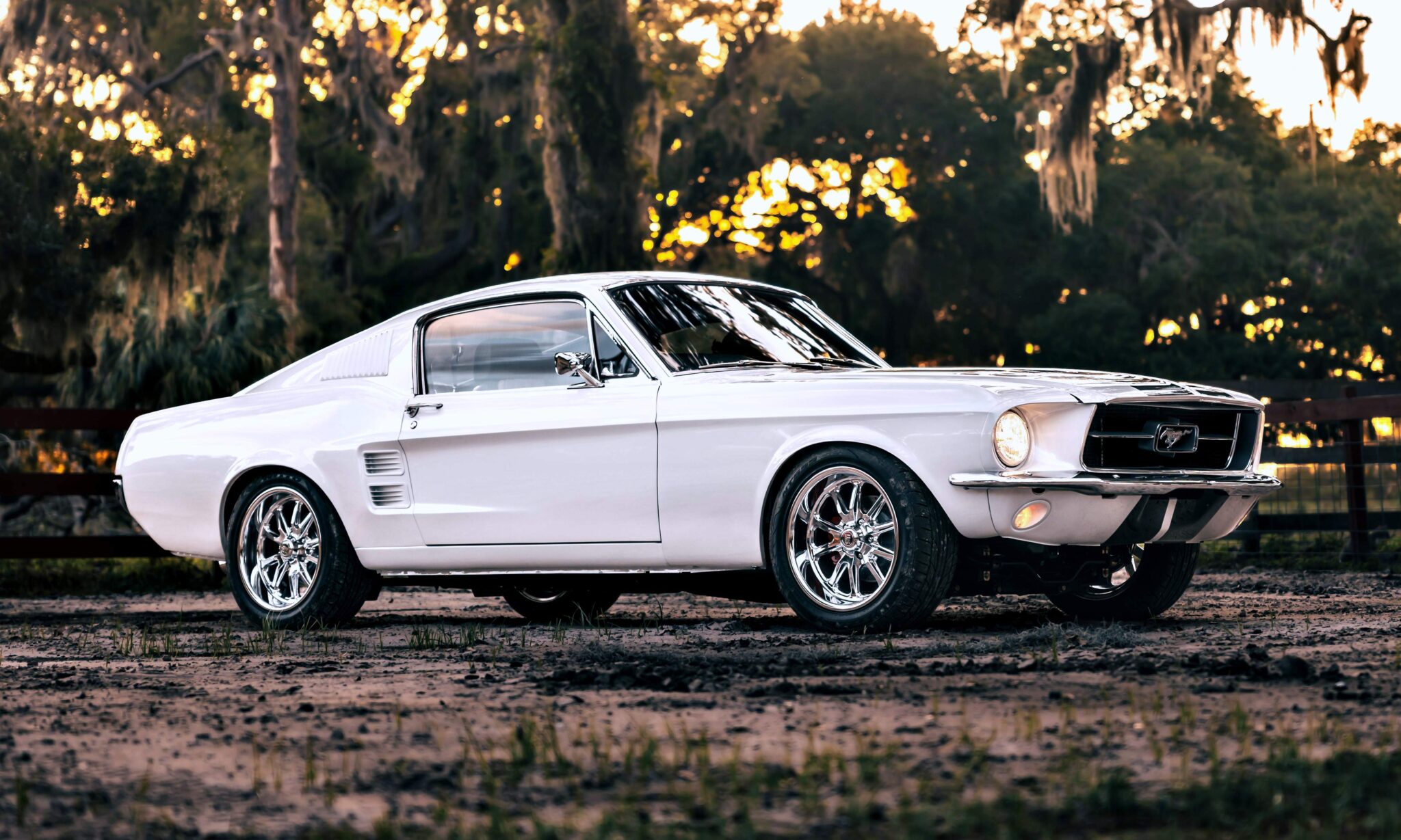 A classic white Ghost Mustang Fastback parked outdoors on a dirt surface, with trees and a wooden fence in the background—a nod to modern muscle and Roush Performance heritage.