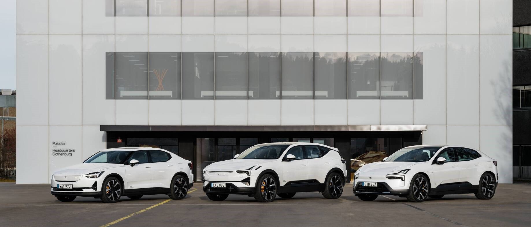 Three white electric cars are parked in front of a modern building with large glass windows.