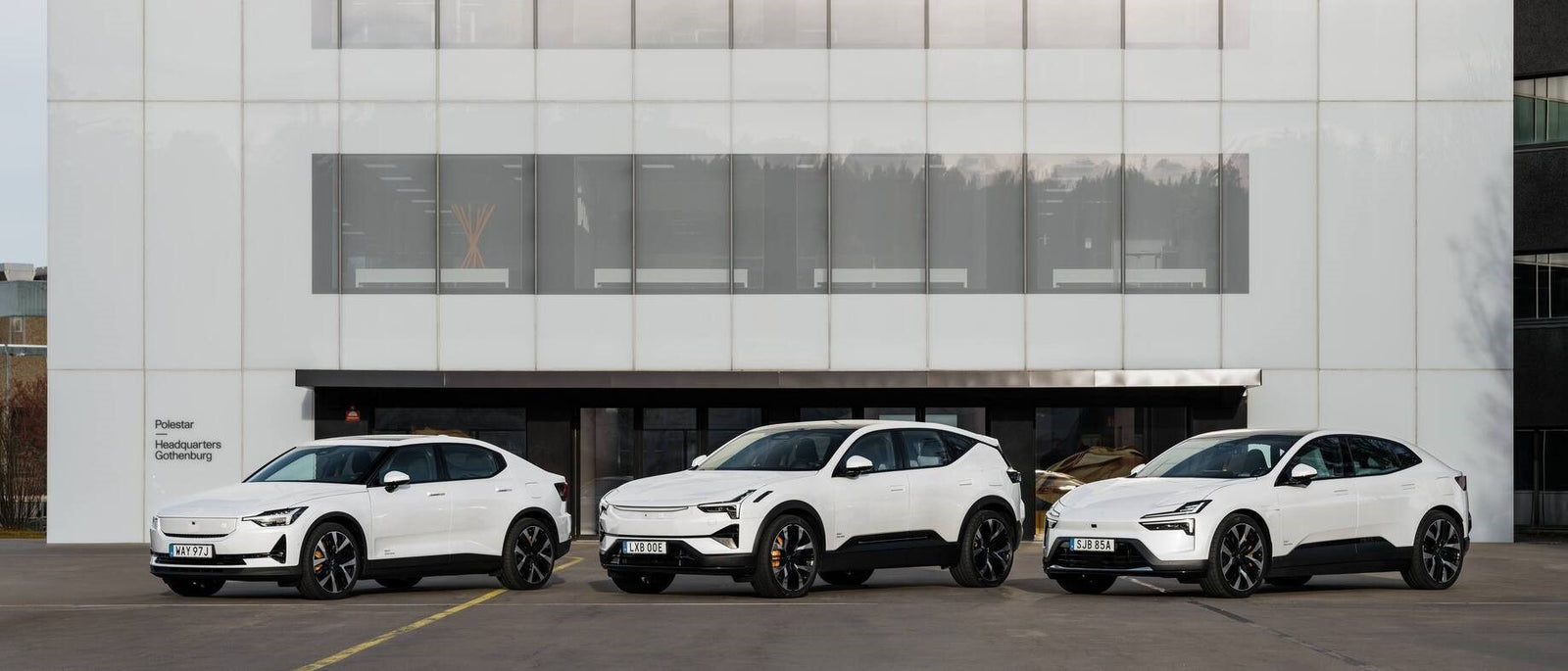 Three white electric cars are parked in front of a modern building with large glass windows.