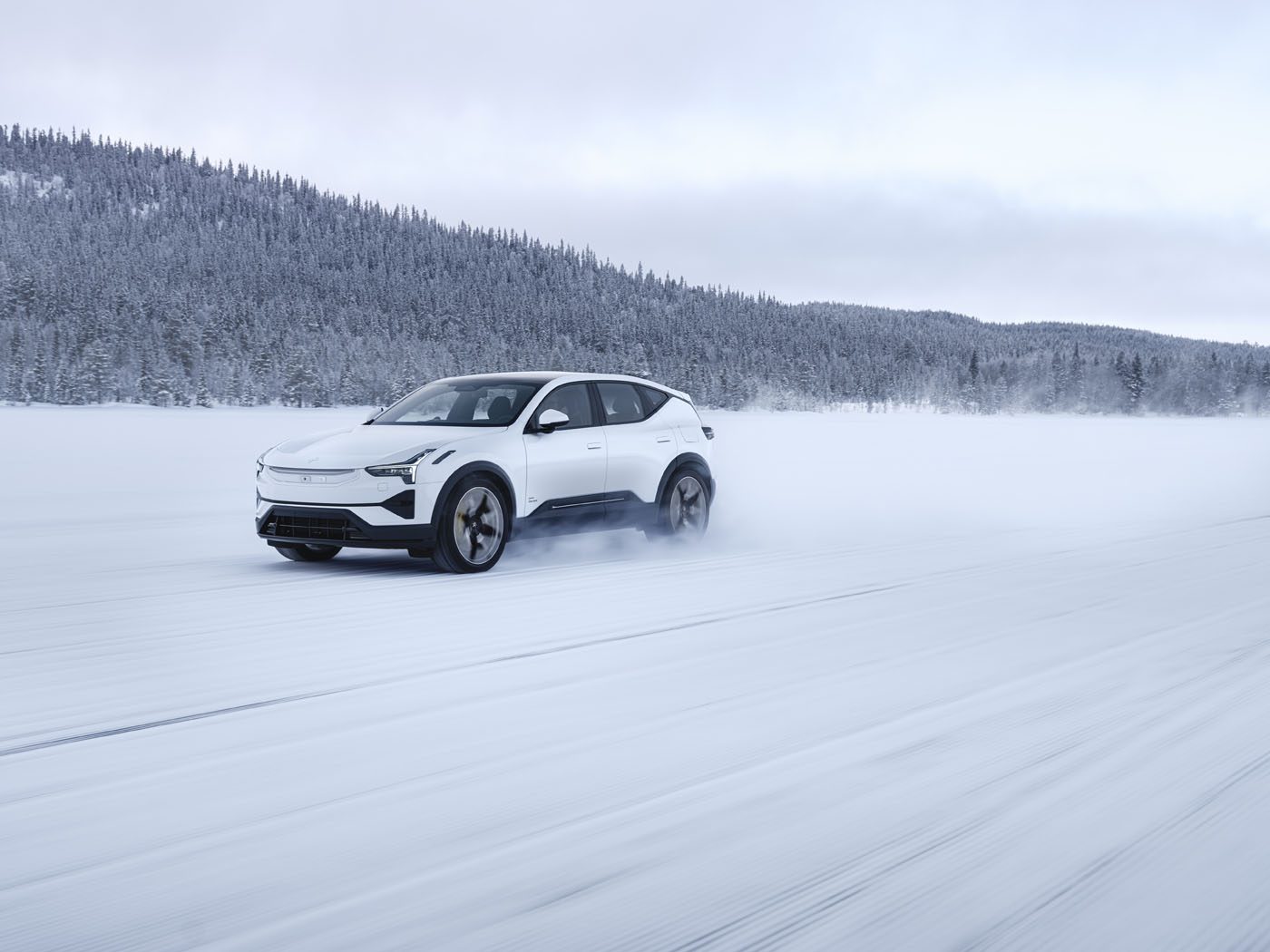 A white SUV drives quickly across a snowy landscape with snow-covered trees in the background.