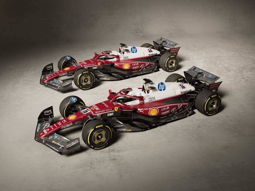 Two Formula 1 cars with red, white, and black livery are parked side by side on a concrete floor, displaying sponsorship logos including Shell, HP, and IBM.