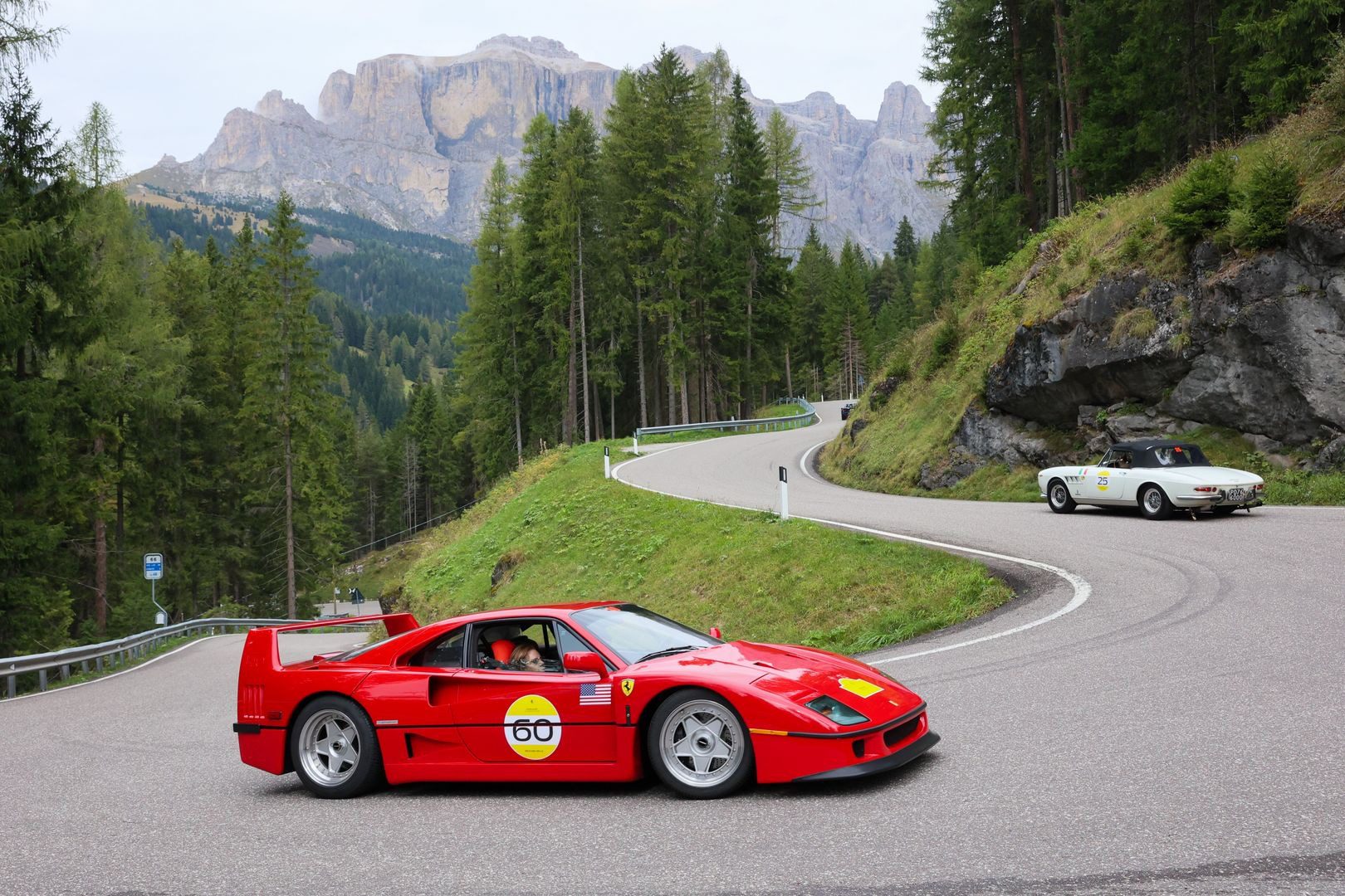 A red Ferrari sports car and a white classic convertible drive on a winding mountain road surrounded by trees and rocky hills.