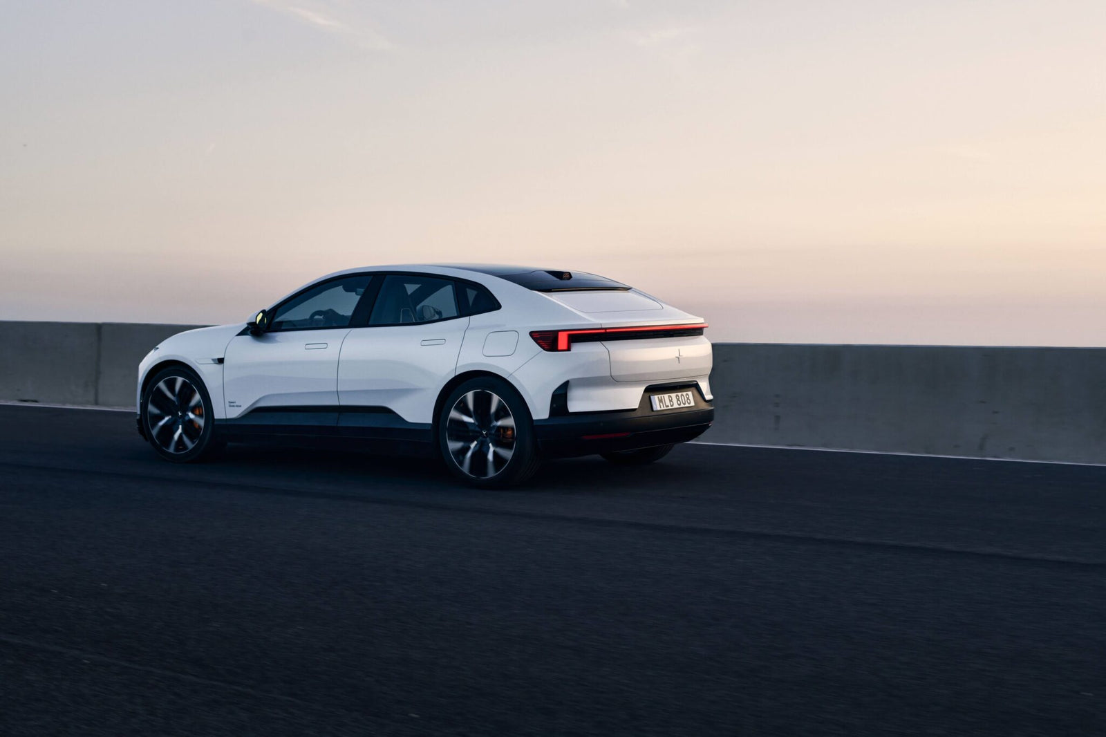 A white electric car drives on an empty road next to a concrete barrier at sunset.