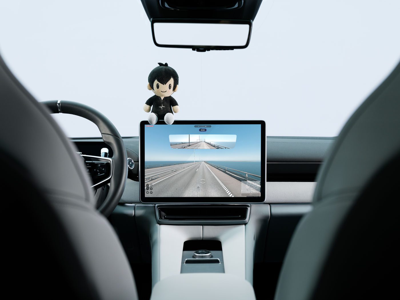 Interior view of a car’s dashboard and steering wheel, with a touchscreen displaying a road—perfect for a GeoGuessr game competition. A doll sits on top of the screen in this unique Polestar Edition cabin.