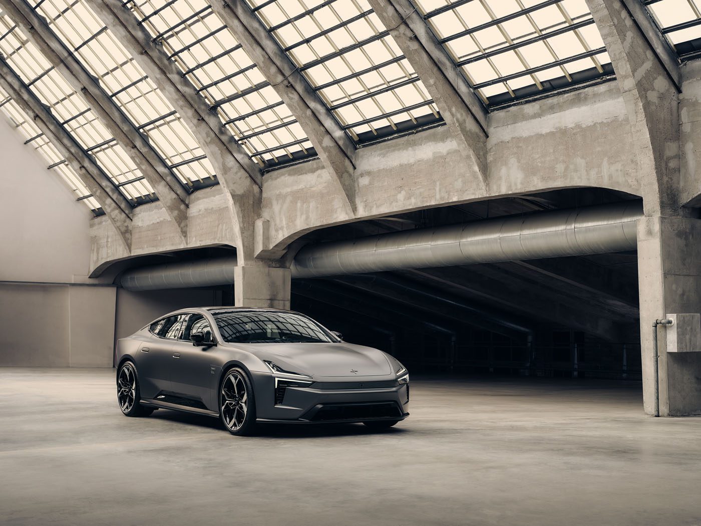 A matte grey Polestar 5 electric sports car is parked in a spacious, industrial-style building with large glass-panel skylights and concrete beams, ready for its cinematic debut in a film starring Alexander Skarsgård.