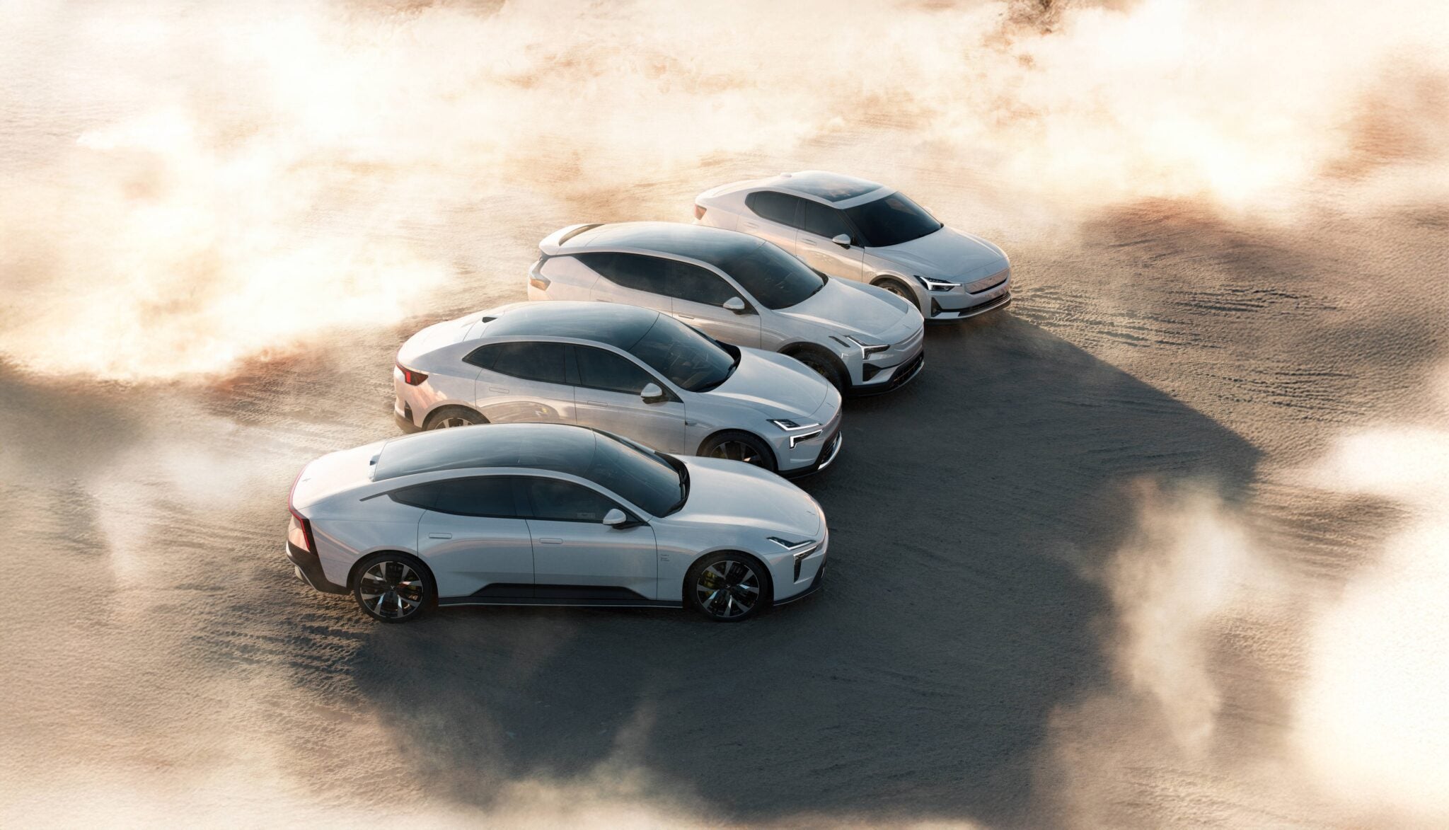 Four white electric cars are parked in a staggered formation on a sandy surface with dust and sunlight surrounding them.