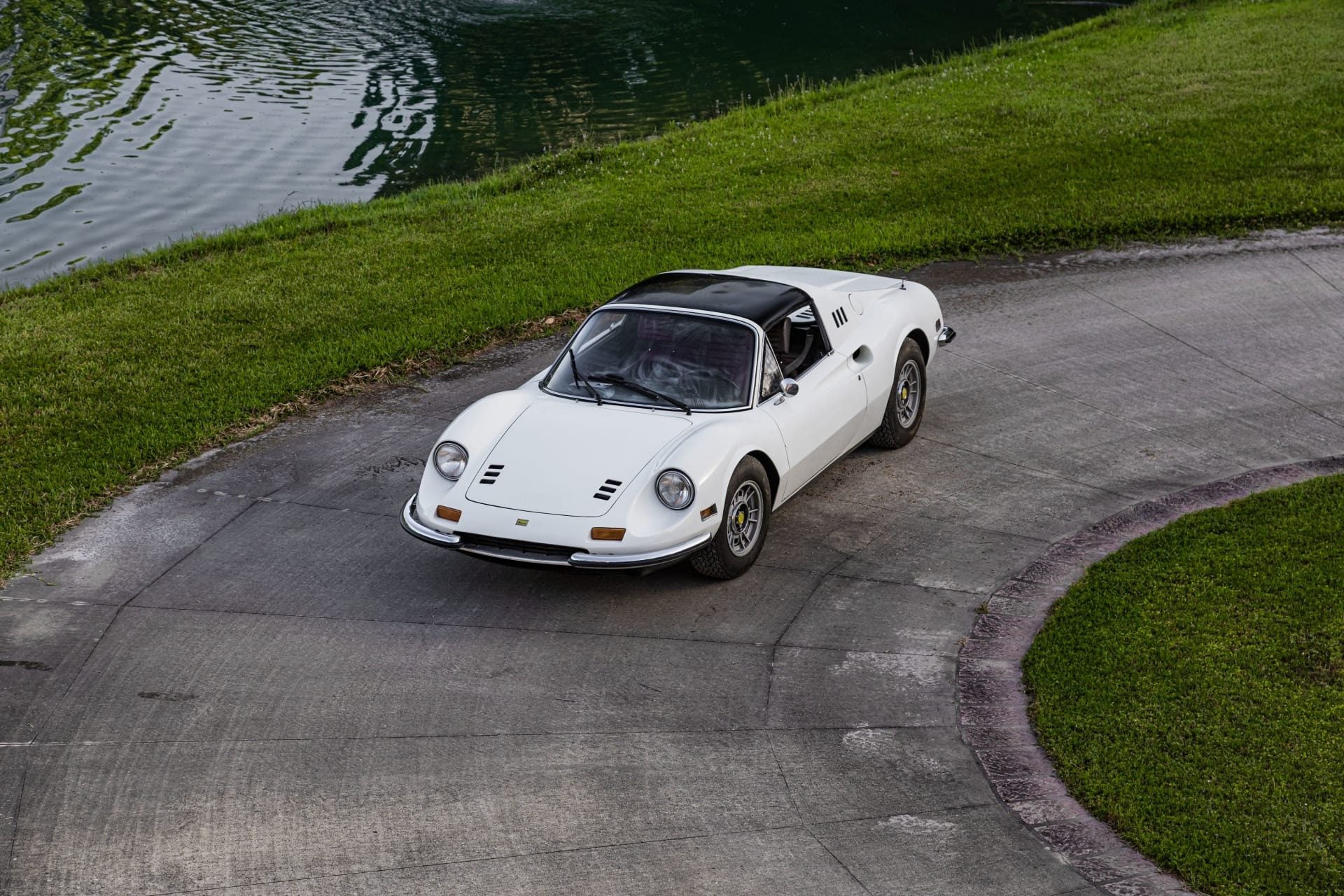 A white classic Ferrari Dino sports car with a black roof is parked on a curved concrete driveway next to a grassy area and a pond, evoking the spirit of the Cannonball Run.