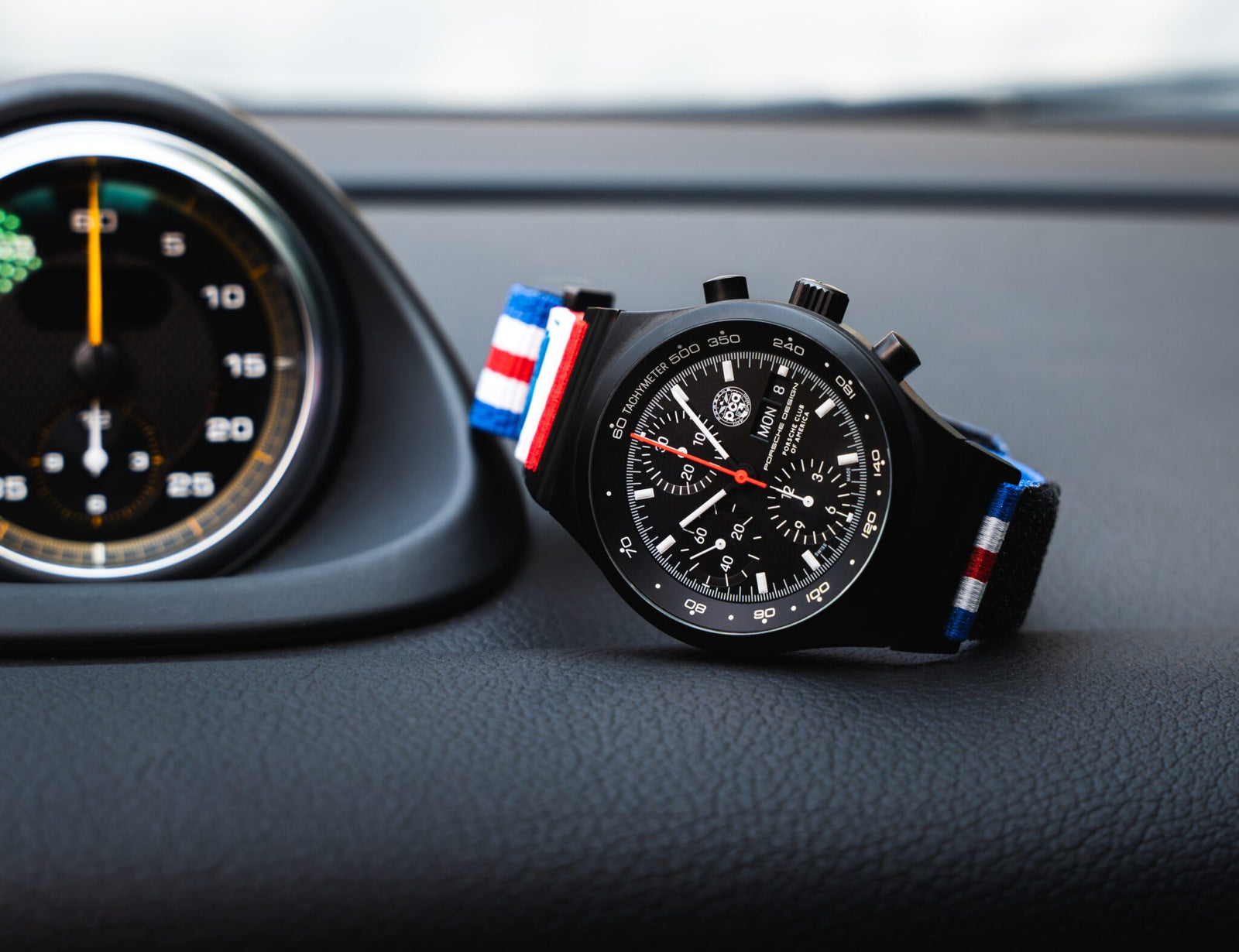 A black Chronograph 1 wristwatch with a red, white, and blue striped fabric strap, designed by Porsche Design, displayed on a car dashboard next to a round speedometer—perfect for PCA enthusiasts.