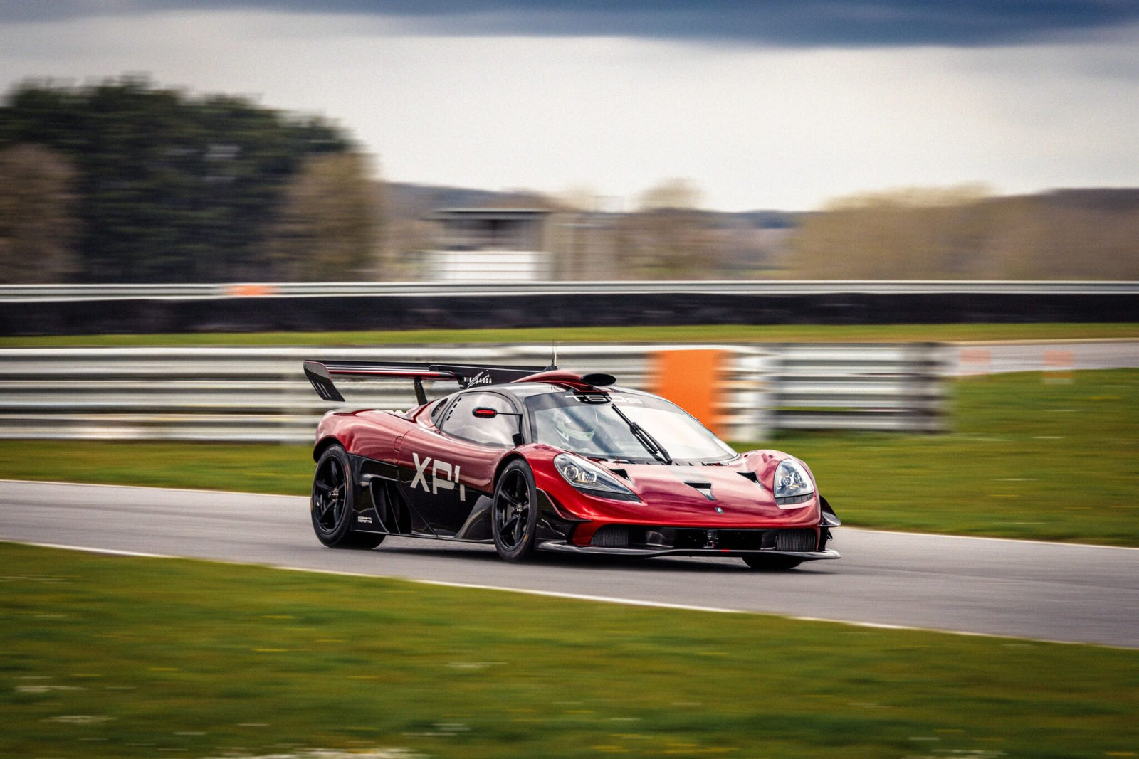 A red sports car with a large rear wing speeds on a racetrack, blurring the background of grass and trees.