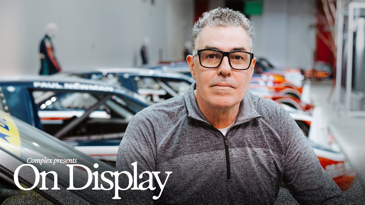 A man wearing glasses and a gray zip-up shirt stands in front of a row of cars in an indoor setting, reminiscent of the Paul Newman car collection. Text on the image reads "Complex presents On Display.