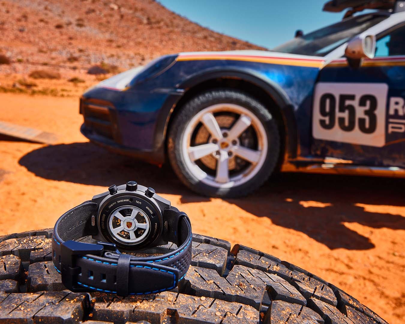A sleek wristwatch with a black band rests on a tire in the desert, set against the backdrop of a race car labeled "953," capturing the essence of Porsche Design.