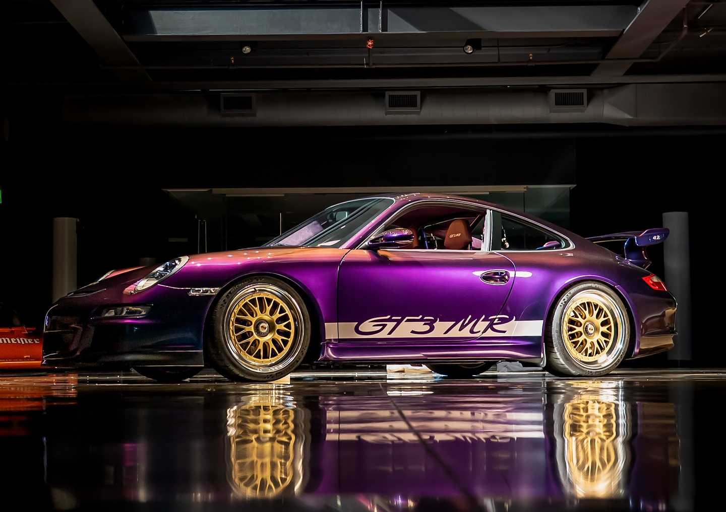 A purple Porsche 997 GT3 MR with gold wheels is parked indoors on a reflective floor under bright lighting, reminiscent of the Werks Reunion Monterey scene.