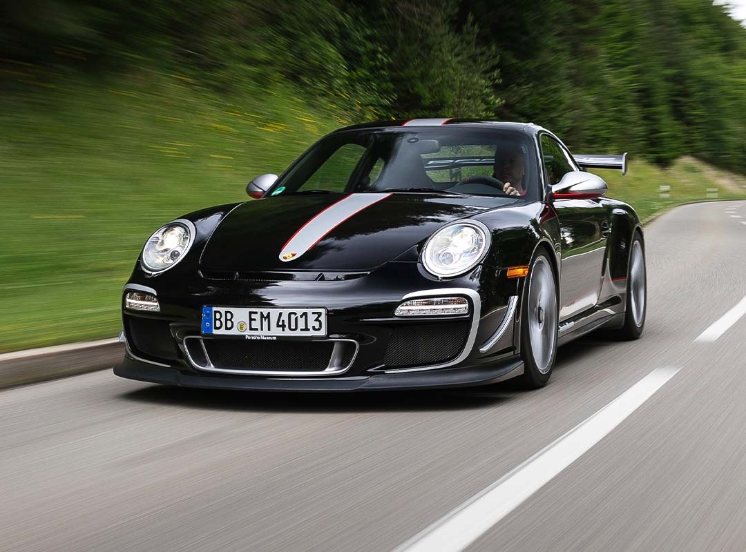 A black 997 Porsche 911 GT3 RS 4.0 with red and white stripes drives on a winding road bordered by greenery and trees.