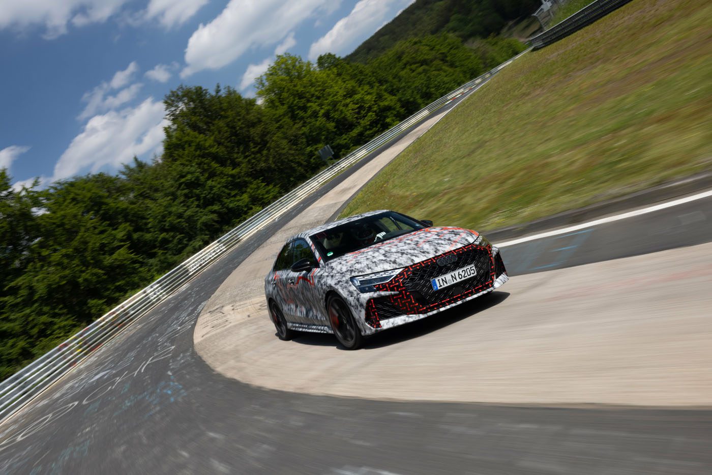 A camouflaged car with German license plates drives through a curve on a racetrack surrounded by grass and trees under a partly cloudy sky.