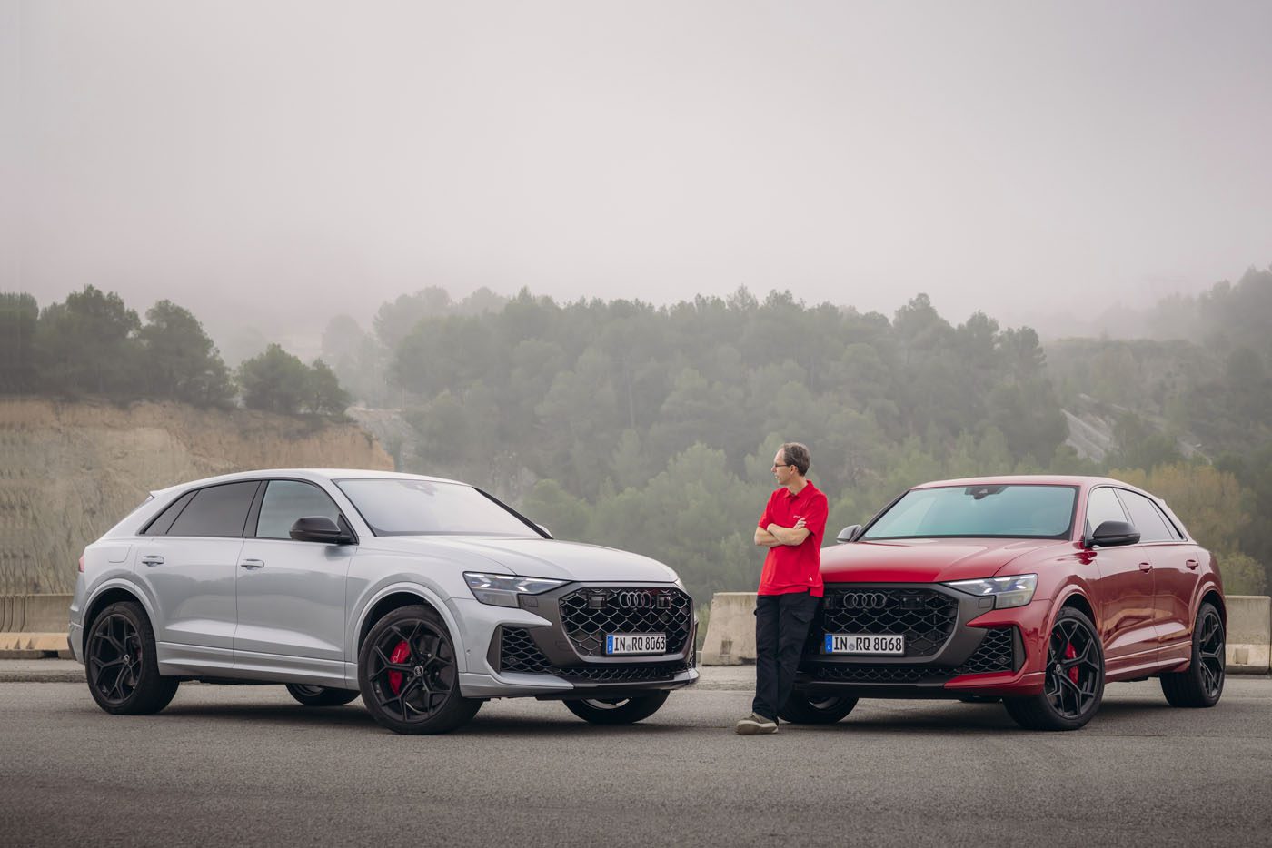 A person, possibly a chassis developer, stands next to two parked Audi SUVs—an elegant gray and a striking red—on a foggy Spanish road. Each vehicle exudes the performance prowess of the Audi RS Q8.