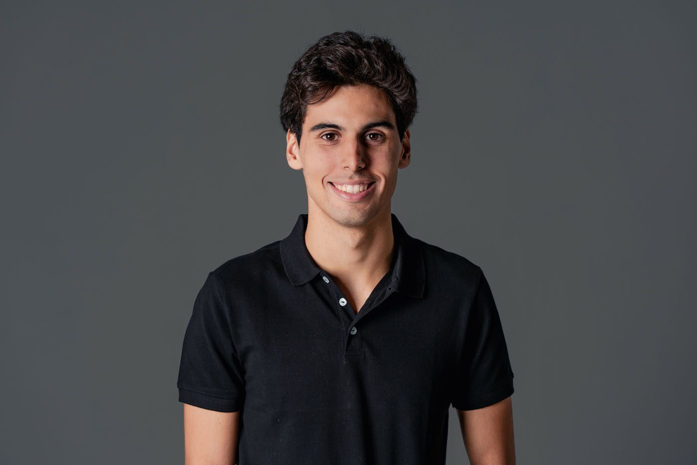 A person with short dark hair, reminiscent of F1 Driver Gabriel Bortoleto, stands against a gray background in a black polo shirt, smiling confidently.