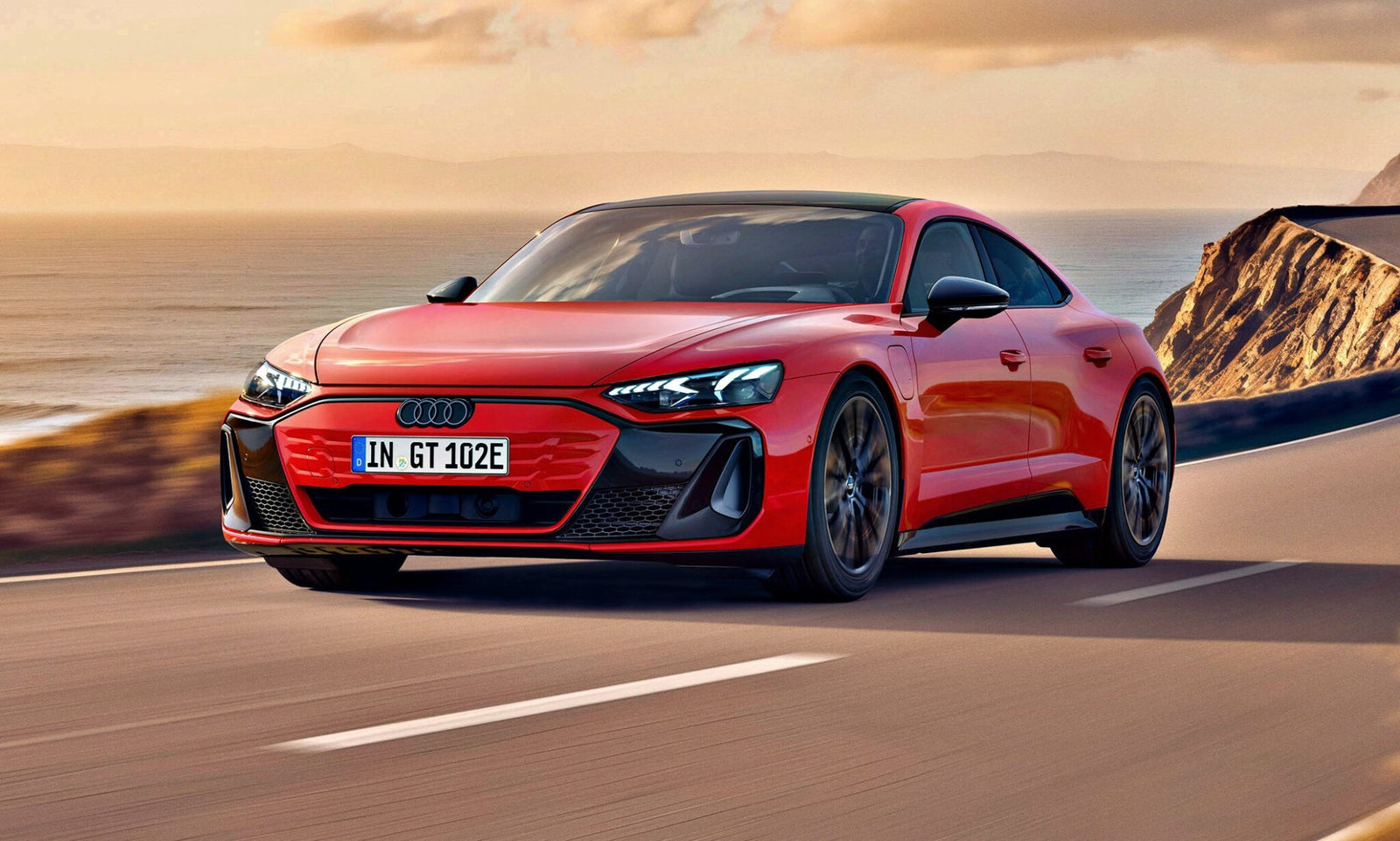 A red Audi electric sports car, inspired by the Most Affordable Audi e-tron GT Debuts And Its Anything But Basic, drives along a coastal road with cliffs and the ocean visible in the background under a partly cloudy sky.