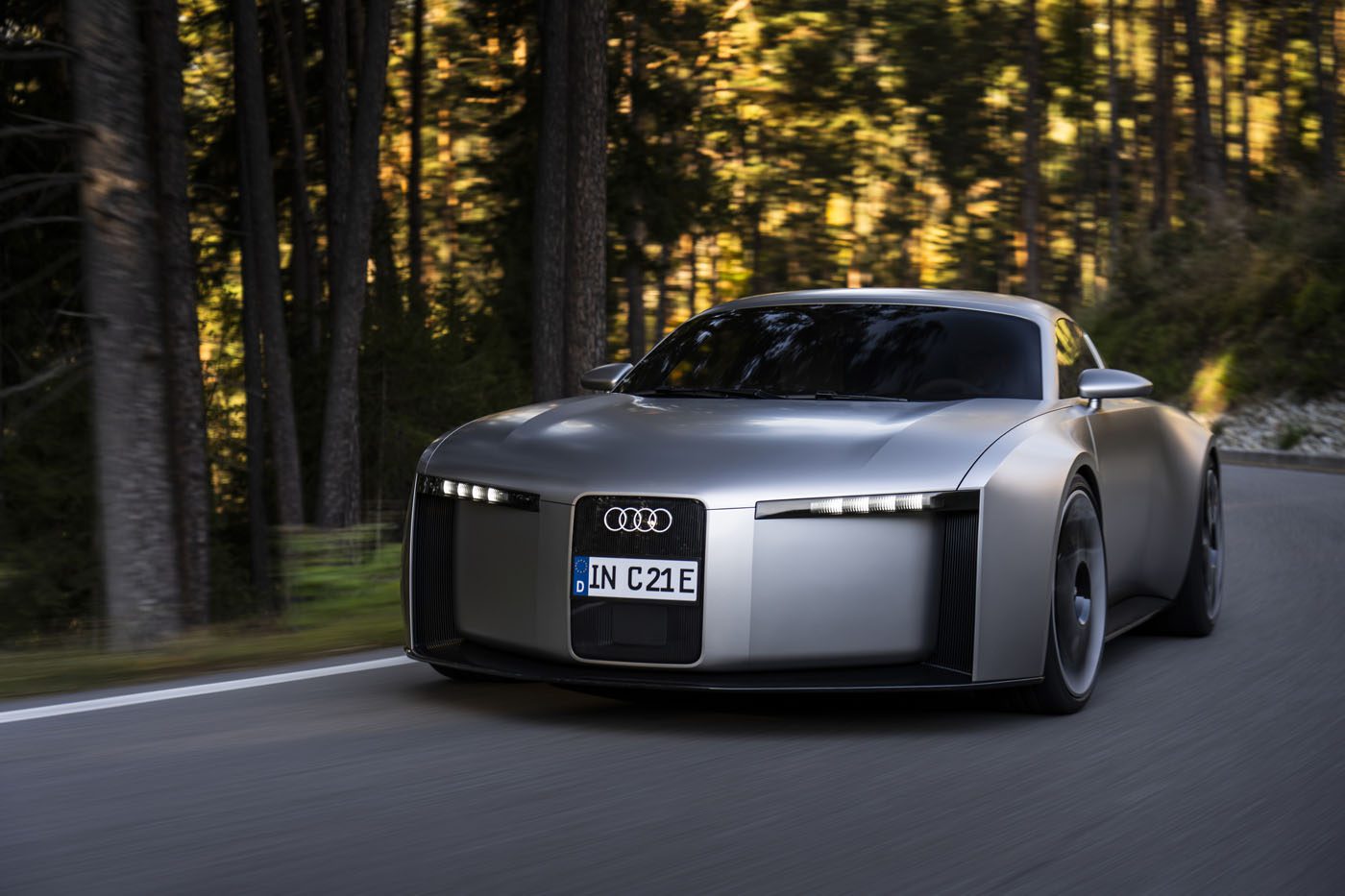A silver Audi Concept C, a street-legal production sports EV, drives on a forested road during daylight, showcasing its modern, minimalist design and illuminated headlights.