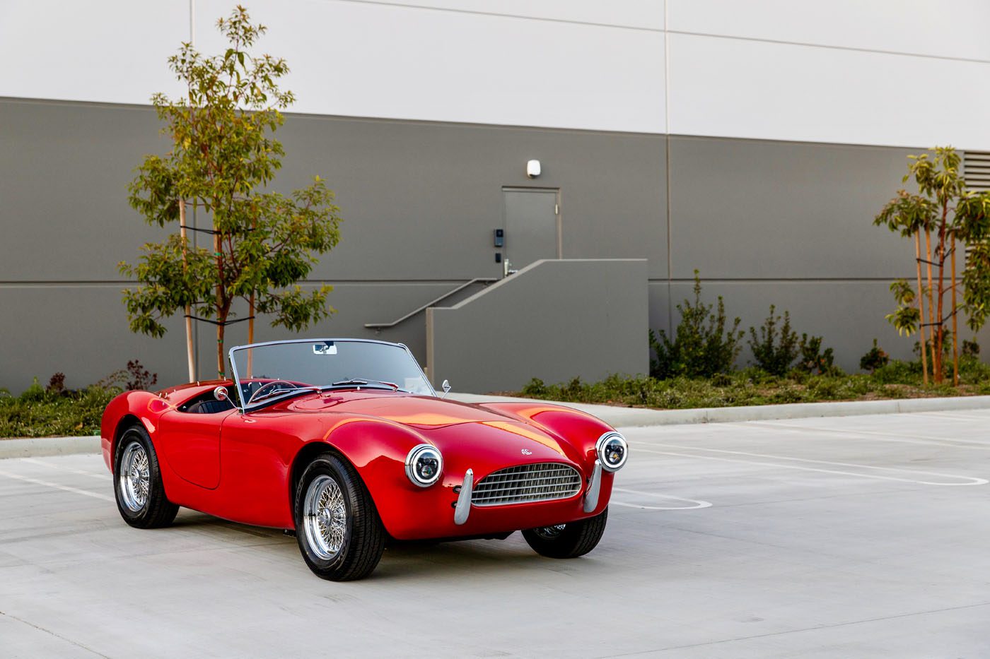 The red, 1-of-49 vintage convertible car from AC Cars sits elegantly in a modern, empty parking lot near a building with trees nearby.